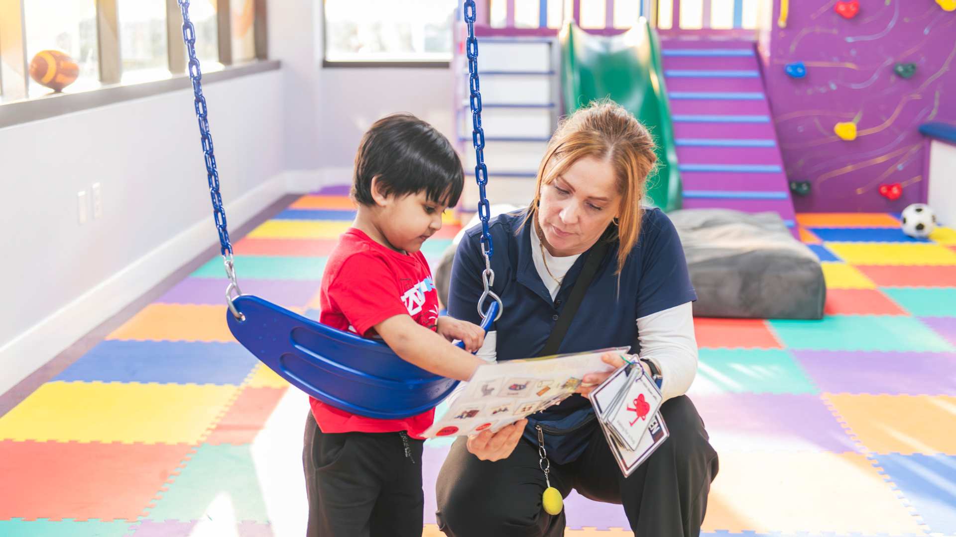 An adult reading a picture book with a child on a swing in a colorful indoor therapy space