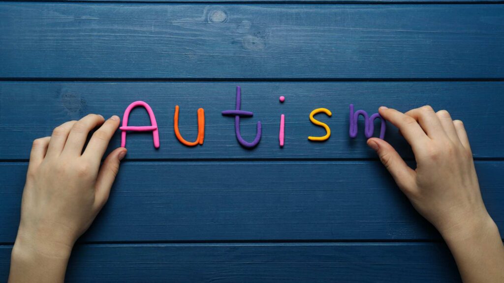 Hands arranging colorful letters spelling the word autism on a blue wooden surface