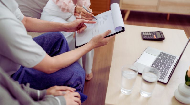 Two individuals reviewing documents on a clipboard in a well-lit, casual setting. One person, wearing a light-colored shirt, points to the paper, while the other, in a pink top, holds a pen. A laptop, calculator, and glasses of water are visible on a wooden table, suggesting a professional or financial discussion regarding health insurance.