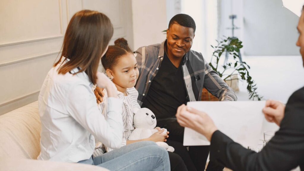 Young girl holding a stuffed animal sits with her parents on a couch while a therapist explain How early should ABA therapy start