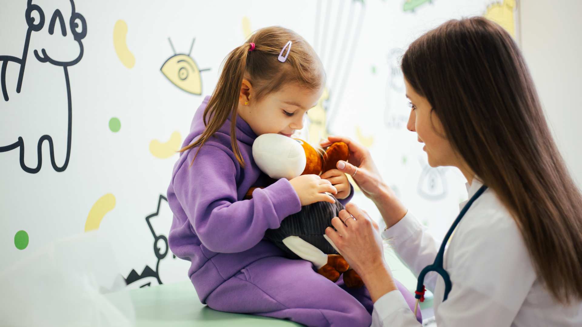 Young girl in a purple hoodie hugs a stuffed animal while a developmental pediatrician examines her in a colorful clinic room